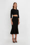 A woman wearing a Victoria Beckham VB Body Cropped Cardigan In Black and a black mermaid skirt stands against a plain white background. The form-fitting silhouette highlights her long hair and high-heeled sandals, exuding a feminine sensibility.