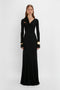 A woman in a long-sleeve black gown with a twist-front design, accessorized with contemporary accessories including Victoria Beckham's Exclusive Perfume Cuff In Gold earrings and bracelets, standing against a white background.