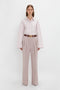 A person stands against a plain white background, wearing a light-colored Button Detail Cropped Shirt In Rose Quartz by Victoria Beckham and high-waisted, wide-leg trousers with a brown belt. Their arms are relaxed at their sides.