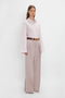 A person is wearing a Victoria Beckham Button Detail Cropped Shirt In Rose Quartz and wide-legged beige trousers with a burgundy belt, standing against a plain white background.