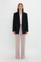 A person stands against a plain white background wearing a black Peak Lapel Jacket In Midnight, light pink shirt, and light pink trousers. With long hair down and hands by their sides, the ensemble exudes a contemporary feel reminiscent of Victoria Beckham's signature style.