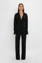 A person stands against a plain white background, wearing a black long-sleeved wrap blouse and sleek Satin Panel Straight Leg Trouser by Victoria Beckham.