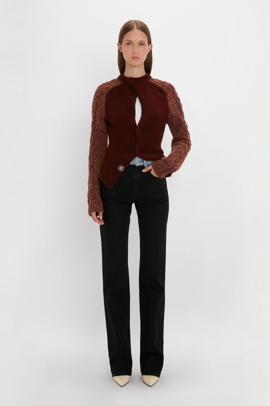 A woman stands against a plain white background, wearing the Victoria Beckham Exclusive Contrast Sleeve Cardigan In Rust, a white top, black pants, and pointed shoes. She has one hand in her pocket and a neutral facial expression.