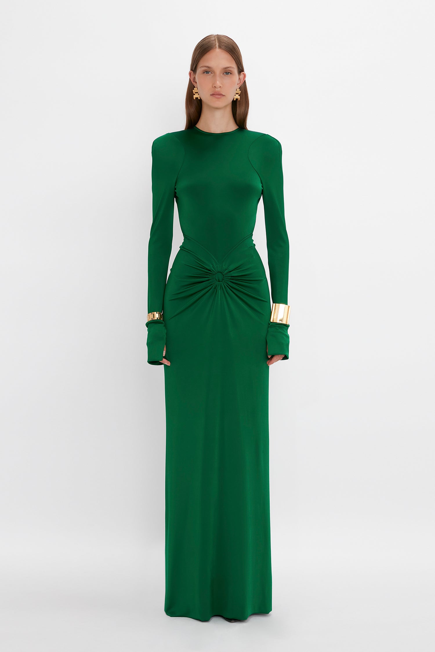 A person stands against a plain white background wearing the Victoria Beckham Circle Detail Open Back Gown In Emerald, a long-sleeved, floor-length green dress made of soft jersey fabric, with a gathered detail at the waist and gold bracelets on both wrists.