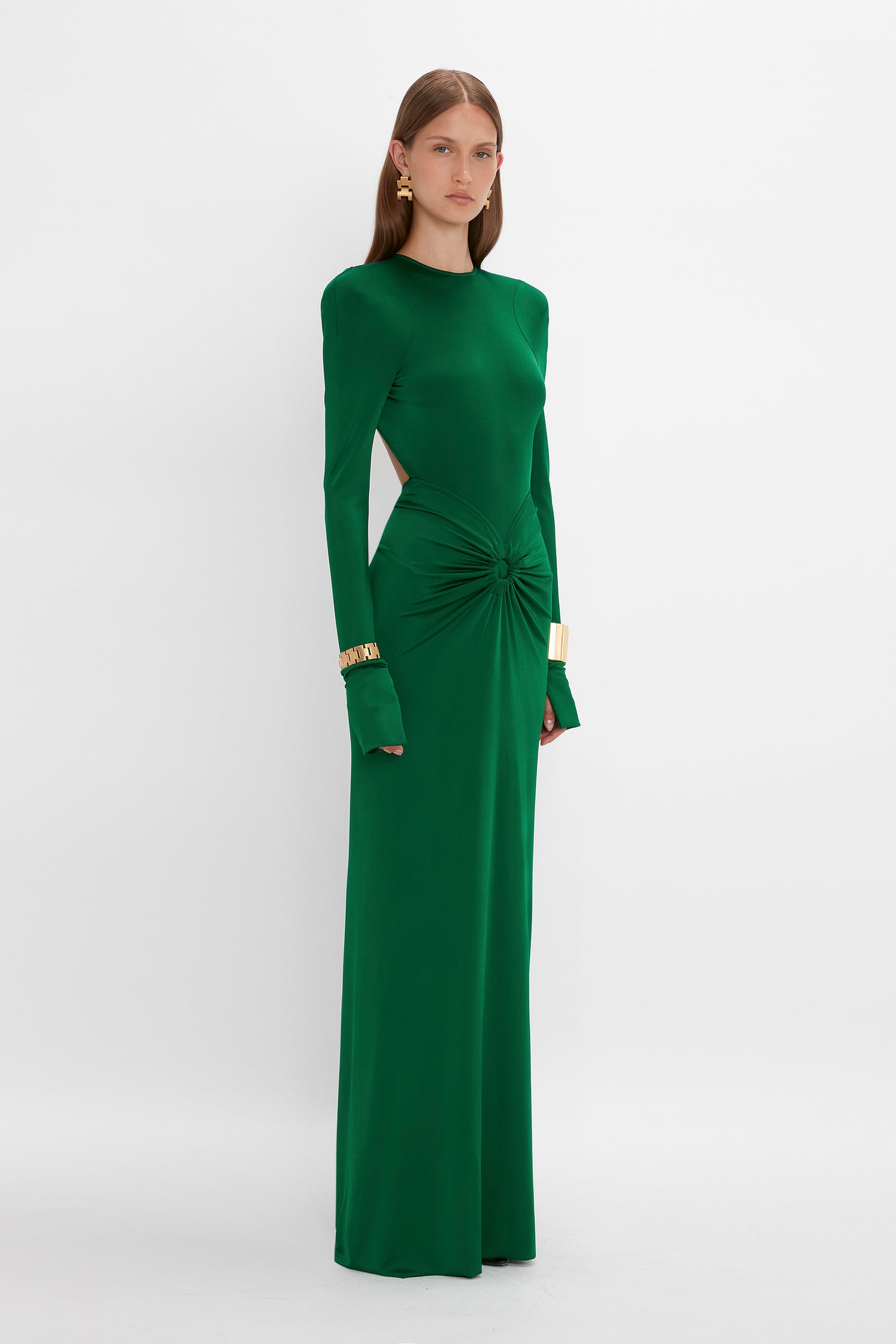 A woman stands wearing a long-sleeved, floor-length green dress with cutout details at the waist and shoulder pads. She is also wearing gold earrings and bracelets. The background is plain white. She is dressed in the Circle Detail Open Back Gown In Emerald by Victoria Beckham.