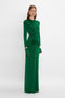 A woman stands wearing a long-sleeved, floor-length green dress with cutout details at the waist and shoulder pads. She is also wearing gold earrings and bracelets. The background is plain white. She is dressed in the Circle Detail Open Back Gown In Emerald by Victoria Beckham.