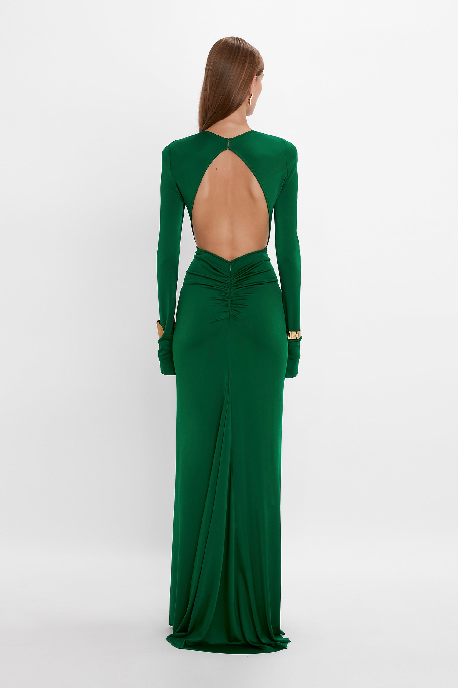 A woman with long hair wearing an elegant, floor-length Victoria Beckham Circle Detail Open Back Gown In Emerald in jersey fabric with an exposed back, stands against a plain white background.