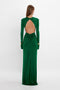 A woman with long hair wearing an elegant, floor-length Victoria Beckham Circle Detail Open Back Gown In Emerald in jersey fabric with an exposed back, stands against a plain white background.