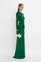 Woman wearing a long-sleeved, green Circle Detail Open Back Gown In Emerald by Victoria Beckham with side cutouts and an exposed back, holding a white clutch and gold accessories, stands against a white background.