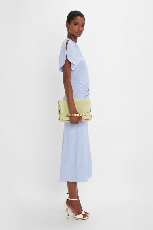 A person stands sideways wearing a light blue dress and white high-heeled sandals, holding an avocado-colored Chain Pouch With Strap In Avocado Leather by Victoria Beckham.