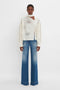 Person wearing a Victoria Beckham Asymmetric Gather Detail Top In Cream paired with faded blue wide-leg jeans, standing against a plain white background.