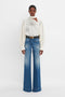 Person standing against a white background, wearing an elegant Asymmetric Gather Detail Top In Cream by Victoria Beckham with blouson sleeves and wide-legged blue jeans with a black belt.