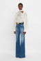 A person stands against a white background wearing an Asymmetric Gather Detail Top In Cream by Victoria Beckham with blouson sleeves and a pair of high-waisted, wide-leg blue jeans.