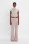 A person stands against a white background, wearing a short-sleeve, high-neck top and high-waisted, wide-leg trousers with a belt. The Victoria Beckham Polo Neck Knitted T-Shirt In Cream adds a sophisticated touch to the ensemble.