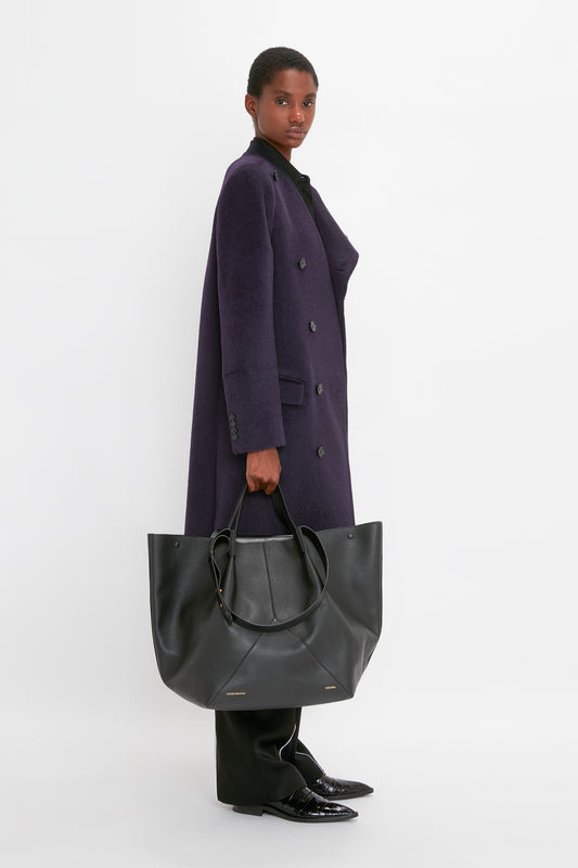 Person in a purple coat holding a Victoria Beckham W11 Jumbo Tote In Black Leather with an internal pocket compartment, standing against a plain white background.