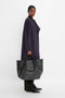 A person stands against a white background wearing a long, dark purple coat, black pants, and black shoes, holding a W11 Jumbo Tote Bag In Black Leather by Victoria Beckham made of luxury grained leather with adjustable straps and an internal pocket compartment by their side.