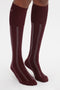 A person's legs are shown wearing knee-high, Victoria Beckham Superfine Rib Socks In Burgundy with vertical stripes on a plain white background.