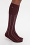 A person is wearing the Victoria Beckham Superfine Rib Socks In Burgundy, which are 100% cotton and feature a long, burgundy and white design with vertical stripes and a small "NB" logo near the top.