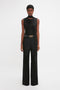 A person stands against a plain background, wearing the Victoria Beckham Sleeveless Tie Neck Top In Black, crafted from luxurious crepe de chine silk. The outfit is paired with high-waisted black wide-leg trousers and a belt.