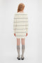 A person with long, light-colored hair is standing with their back to the camera, wearing a cream-colored, long-sleeved, striped **Frame Detail Jumper Dress In Natural-Navy** by **Victoria Beckham**, gray knee-high socks, and black shoes.