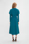 A person with long blonde hair is wearing a teal Long Sleeve Dolman Midi Dress In Petroleum by Victoria Beckham made from fluid cady fabric and black high-heeled sandals, facing away from the camera against a plain white background. The entire ensemble exudes contemporary sophistication.