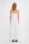 Rear view of a person with wavy, light brown hair, wearing the Victoria Beckham Cami Fit And Flare Midi In White, a white, sleeveless, ankle-length summer dress with adjustable straps and a zipper down the back. Simple white background.