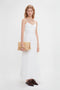 A woman stands against a white background, wearing the Victoria Beckham Cami Fit And Flare Midi In White made of breathable stretch cotton with adjustable straps, and holding a tan clutch. She has light-colored hair and is wearing dark, lace-up shoes.