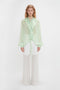 A person stands against a plain white background, adorned in a Romantic Blouse In Jade by Victoria Beckham with diaphanous blouson sleeves, paired with white wide-legged sheer trousers.