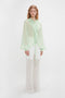 A person stands against a plain background, wearing the Victoria Beckham Romantic Blouse in Jade with diaphanous blouson sleeves and transparent white pants.