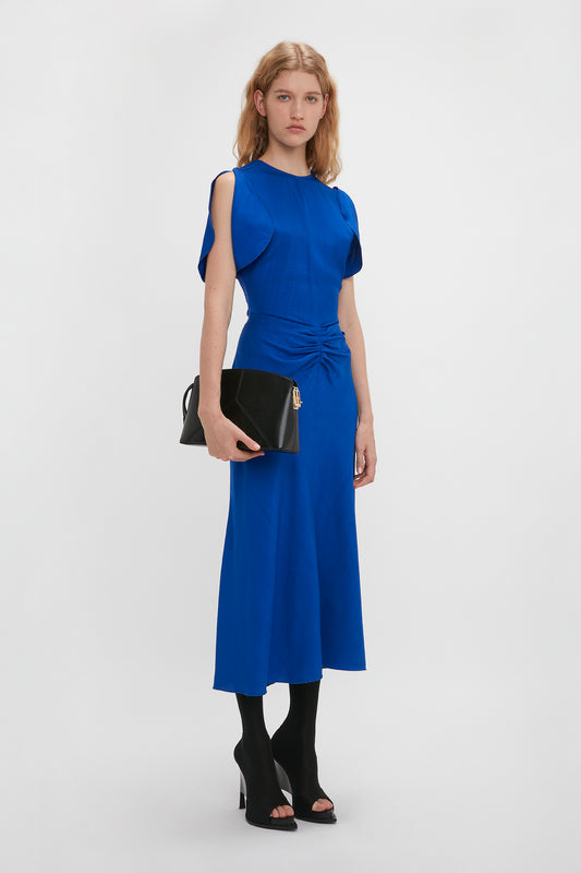 A woman wearing a royal blue dress and black heels holds a Victoria Clutch Bag In Black Leather by Victoria Beckham with a structured silhouette against a plain white background.