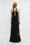 A person with long, wavy hair wearing a floor-length, sleeveless black dress is standing and facing away from the camera. The Sheer Cami Gown In Black-White Contorted Net by Victoria Beckham features a racerback design and a subtle twisted net print on the lower right side.