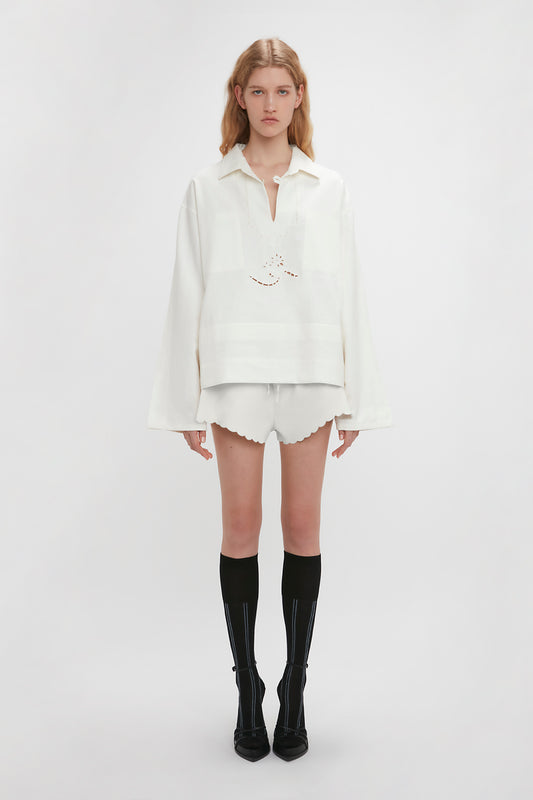 A young woman stands against a white background, wearing the Victoria Beckham Oversized Embroidered Tunic In Antique White and white shorts with scalloped edges, complemented by black knee-high socks with heels.