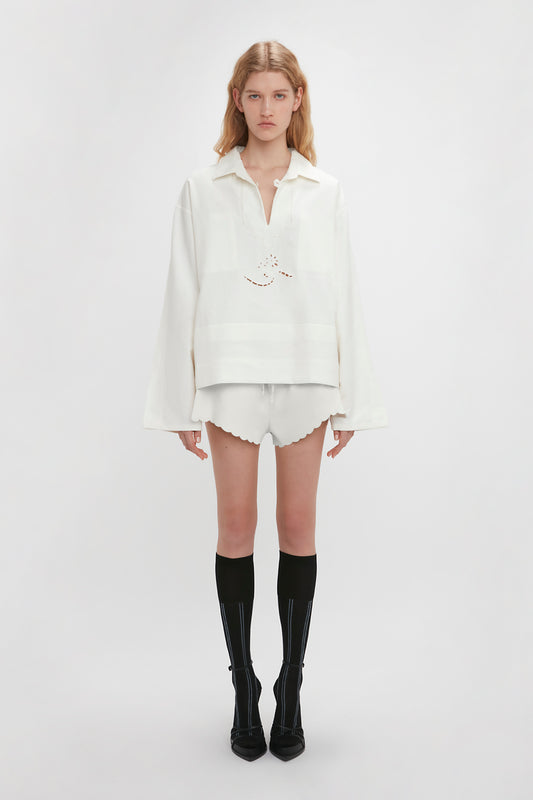 A person stands against a plain white backdrop wearing an antique white long-sleeve top, Victoria Beckham Drawstring Embroidered Mini Short In Antique White, black knee-high socks, and black shoes.