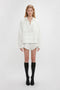 A person stands against a plain white backdrop wearing an antique white long-sleeve top, Victoria Beckham Drawstring Embroidered Mini Short In Antique White, black knee-high socks, and black shoes.