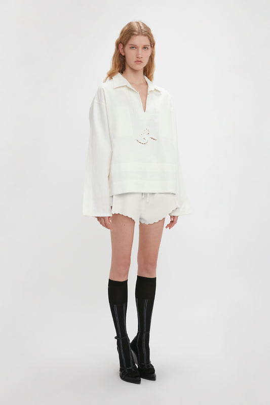A woman stands against a plain background, wearing an Oversized Embroidered Tunic in Antique White by Victoria Beckham, white shorts with a scalloped hem, and black knee-high socks.