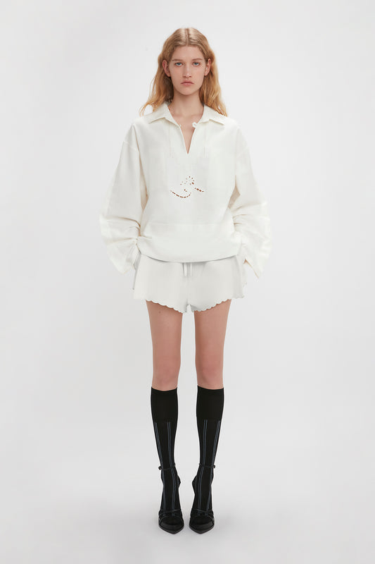 A person stands against a plain background wearing an Antique White pullover, Victoria Beckham Drawstring Embroidered Mini Short In Antique White, and black knee-high socks with black pointed shoes. Their hands are down by their sides.