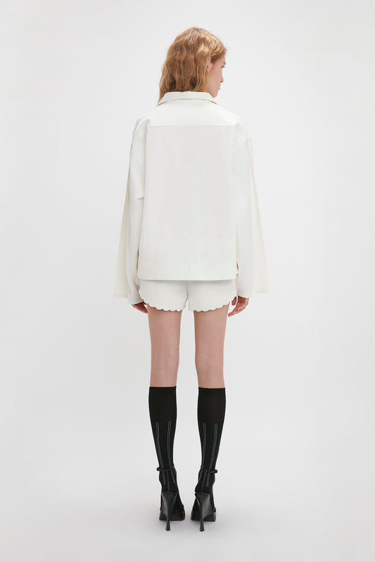 Woman standing with her back to the camera, wearing a white long-sleeve jacket with embroidered detailing, Victoria Beckham Drawstring Embroidered Mini Short In Antique White, black knee-high socks, and black high-heeled shoes against a plain white background.