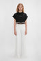 A person with long hair stands against a plain background, wearing a black 'Do As I Say, Not As I Do' Slogan T-Shirt In Black by Victoria Beckham and white wide-legged pants, along with a brown belt.