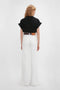 Woman standing with her back to the camera, wearing a black 'Do As I Say, Not As I Do' Slogan T-Shirt by Victoria Beckham and white wide-legged pants with a brown belt.