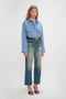 A person stands against a plain background wearing a Cropped Seam Detail Shirt In Steel Blue by Victoria Beckham, faded blue jeans, a brown belt, and black high-heeled sandals.