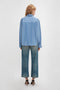 Person with long hair wearing a Victoria Beckham Cropped Seam Detail Shirt In Steel Blue made from organic cotton, jeans, and black heels, standing and facing away from the camera in a white studio setting.