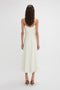 A woman with wavy, brown hair stands with her back to the camera, wearing a white, sleeveless, ankle-length Victoria Beckham Exclusive Panelled Midi Cami Dress In Vanilla featuring a square neckline and brown high-heeled shoes against a plain white background.