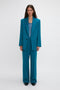 A person with long hair is standing straight, showcasing an oversized silhouette in a teal suit with a Peak Lapel Jacket In Petroleum from the Victoria Beckham brand, paired with a matching teal top and black shoes, against a plain white background.