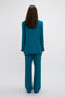 A person with long hair is standing facing away from the camera, wearing a teal suit with an oversized silhouette featuring a Peak Lapel Jacket In Petroleum by Victoria Beckham and wide-legged pants against a plain background.