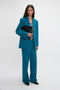 A person with long hair wearing an oversized teal suit, featuring the Waistband Detail Straight Leg Trouser In Petroleum by Victoria Beckham, holds a large black clutch while standing against a plain white background.