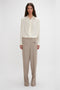 A person stands against a plain background wearing an Asymmetric Ruffle Blouse In Ivory by Victoria Beckham, featuring voluminous sleeves, and beige trousers.