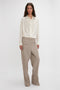 Person standing against a plain background, wearing Victoria Beckham's Asymmetric Ruffle Blouse In Ivory and wide-legged beige trousers.