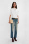 A person stands in a relaxed cut Bonded Jersey Tee In White by Victoria Beckham, blue jeans, and dark shoes, holding a beige clutch bag. The plain white background adds to the simplicity.