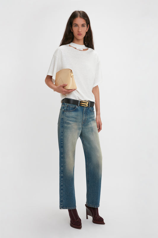 A woman with long hair wears a white t-shirt, blue jeans, and maroon heels. She stands against a plain white background, holding an elegant Victoria Clutch Bag In Sesame Leather from Victoria Beckham.