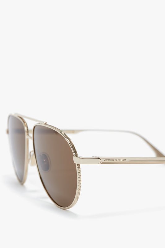 Close-up of V Metal Pilot Sunglasses In Gold-Khaki with brown lenses and adjustable nose pads on a white background. The product has the "Victoria Beckham" logo inscribed on the arm.